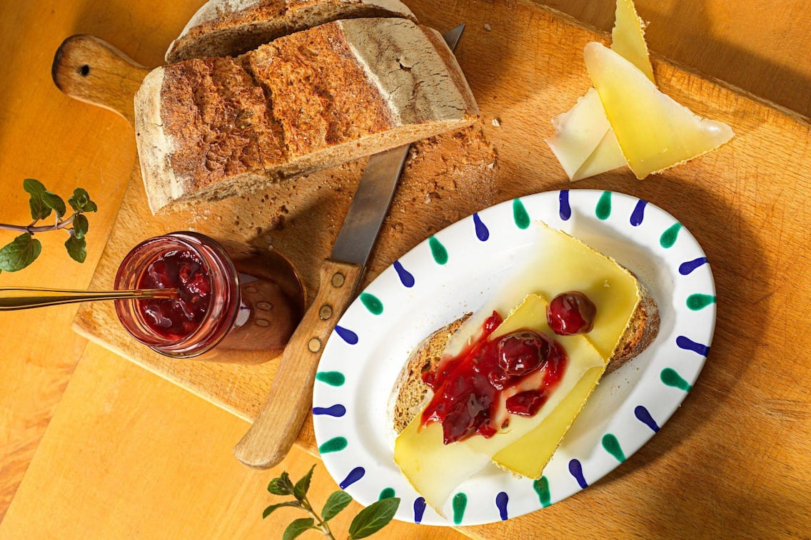 Auf einem Holzbrett steht ein halber Brotlaib ein offenen Kirschmarlmeladeglas mit einem Löffel drin, danaben steht ein ovals Gmundner Teller mit einem Stück Brot, Käsescheiben und Kirschmarmelade oben drauf.