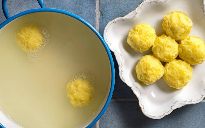 Blauer Email-Kochtopf in dem Kochwasser mit zwei Waldviertler Erdäpfelknödel schwimmen. Daneben steht eine Schüssel mit geschwungenem Goldrand und darin liegen sechs weiter Erdäpfelknödel