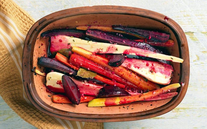 Rote Rüben, gelbe Rüben, weiße Rüben der länge nacht geviertelt in einem Römertopf mit Gewürzen. Daneben liegt ein Geschirrtuch.