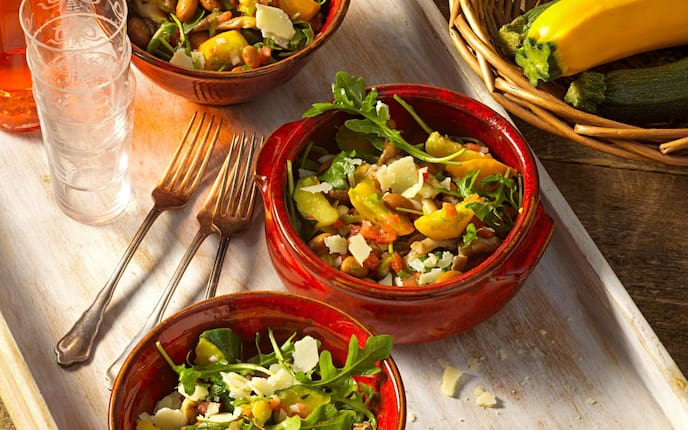 Frischer Salat mit Zucchini, Bohnen, Pilzen und Käse in drei roten Suppenschüsseln angerichtet. Alles steht auf einem Holztablett, mit drei Gabeln, drei ineinandergestapelten Trinkgläsern mit Gravur. Das Tablett steht auf einem alten Holztisch, daneben ist der Anschnitt eines kleinen Brotkorbes zu sehen, in dem zwei grüne und eine gelbe Zucchini liegt.