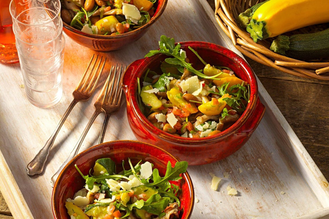 Frischer Salat mit Zucchini, Bohnen, Pilzen und Käse in drei roten Suppenschüsseln angerichtet. Alles steht auf einem Holztablett, mit drei Gabeln, drei ineinandergestapelten Trinkgläsern mit Gravur. Das Tablett steht auf einem alten Holztisch, daneben ist der Anschnitt eines kleinen Brotkorbes zu sehen, in dem zwei grüne und eine gelbe Zucchini liegt.