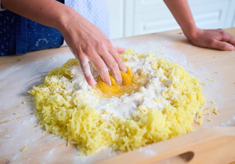Gekochte Erdäpfel, die durch eine Presse gedrückt wurden, liegen auf einem Haufen auf einem Holznudelbrett, in der Mitte mit einer Mulde. Darin sind Mehl und ein Eidotter zu sehen und eine Hand beginnt alles zu vermengen.