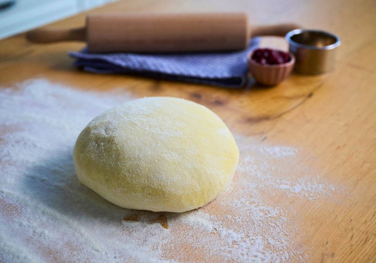 Eine bemehlte glatte Teigkugel liegt auf einem bemehlten Holzuntergrund, daneben sieht man verschwommen ein Nudelholz und blaues Geschirrtuch, einen runden Metallausstecher und ein kleines Schüsserl mit Marmelade.