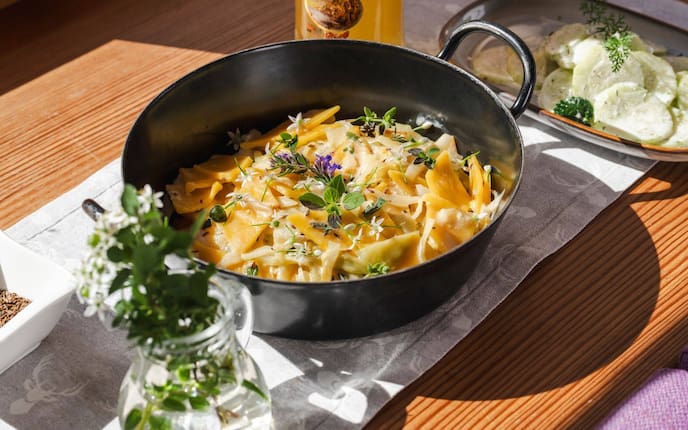 Krautfleckerl in einer Eisenservierpfanne, steht auf einem Holztisch, daneben steht ein Krügerl Bier, und eine kleine Keramikplatte mit Rahmgurkensalat.