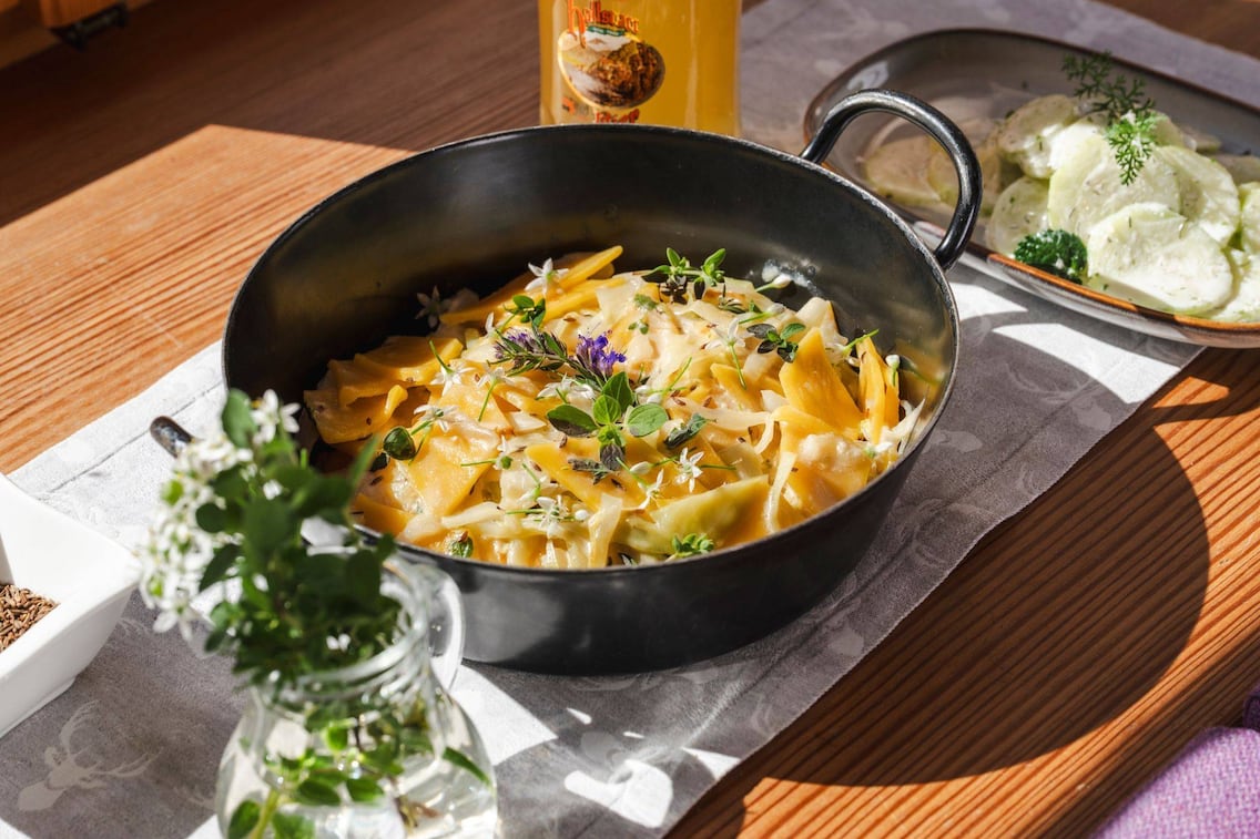 Krautfleckerl in einer Eisenservierpfanne, steht auf einem Holztisch, daneben steht ein Krügerl Bier, und eine kleine Keramikplatte mit Rahmgurkensalat.