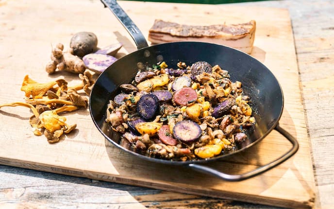 Ein Holztisch im freien auf der ein Holzbrett liegt. Darauf steht eine Eisenpfanne mit einem Erdäpfelgröstel mit Eierschwammerln, violetten, rosa und gelben Erdäpfelscheiben und Speck. Neben der Pfanne liegen violette Erdäpfel, frische Eieschwammer und ein Stück Speck.