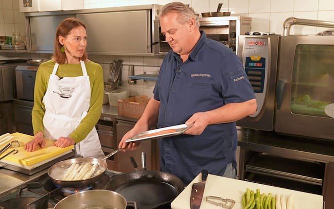 Doris Ahornegger und Richard Poppmeier stehen in seiner Küche und plaudern, während er Spargel in einer Pfanne brät und ein Fischfilet auf einem Teller in der anderen Hand hält.