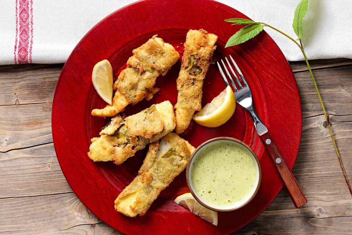 Selbst panierter Fisch mit Zitronenspalten und einer Sauce zum Tippen plus Gabel mit Holzgriff auf einem roten Teller. Alles steht auf einem Holztisch und daneben liegt eine Tischdecke in natur mit roter Bordüre.