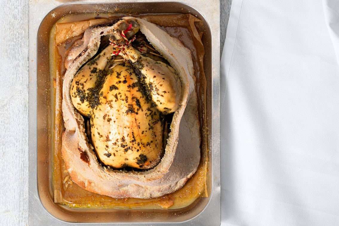 Ganzes Huhn mit Kräutern in einer Alupfanne in Salzteig geschmort, der Salzteig ist aufgeschnitte und man sieht die ganze Henne