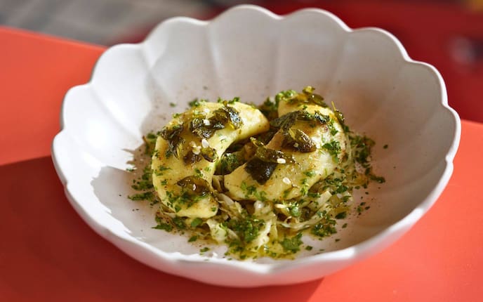 Zwei Teigtascherl sind auf Kraut und mit Kräutern in einer gewellten weißen Schüssel auf einem roten Tisch angerichtet.