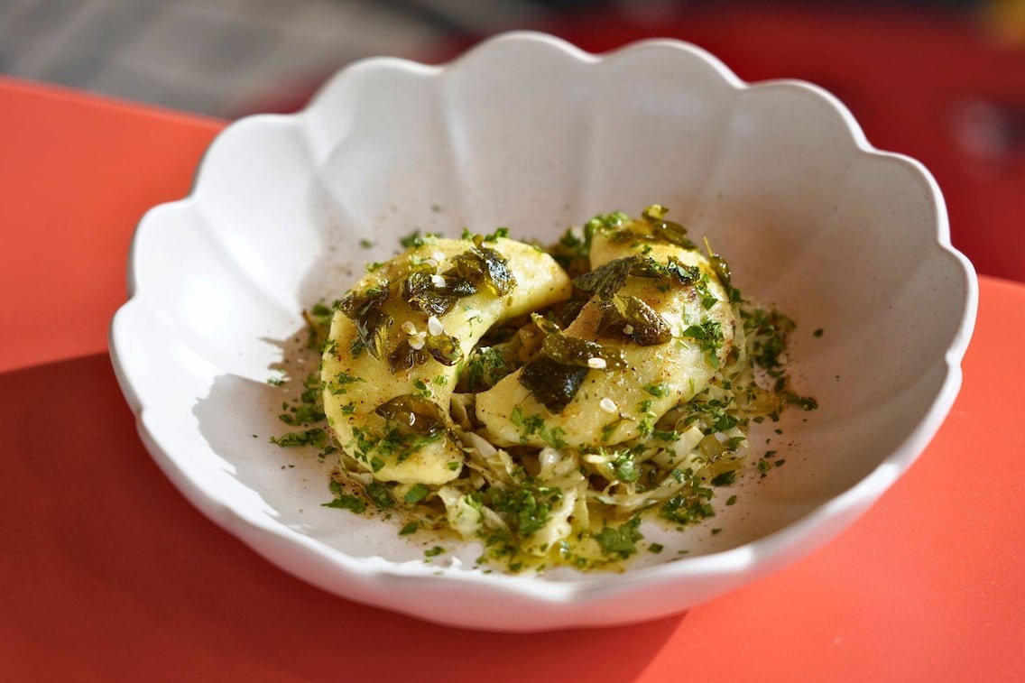 Zwei Teigtascherl sind auf Kraut und mit Kräutern in einer gewellten weißen Schüssel auf einem roten Tisch angerichtet.