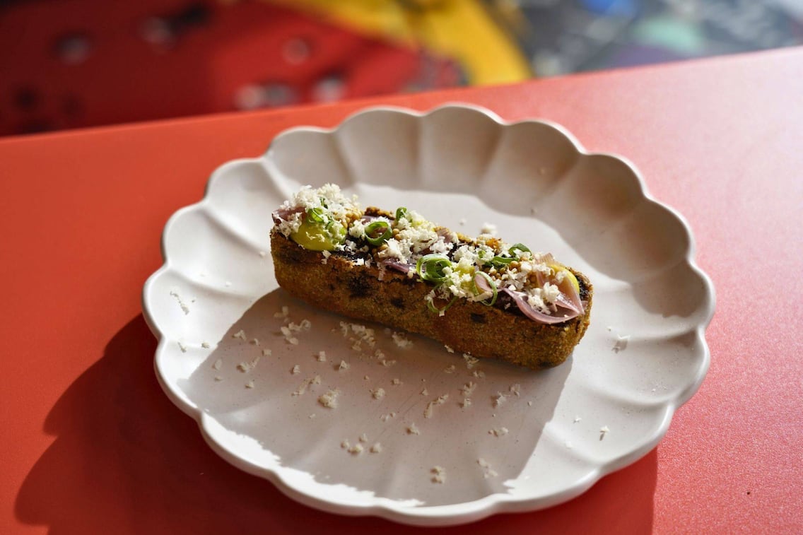 Ein gebackenes Stück Blunze mit Frühlingszwiebelringen, Kren und Perlzwiebel auf einem gewellten weipen Teller angerichtet, der auf einem roten Untergrund steht.