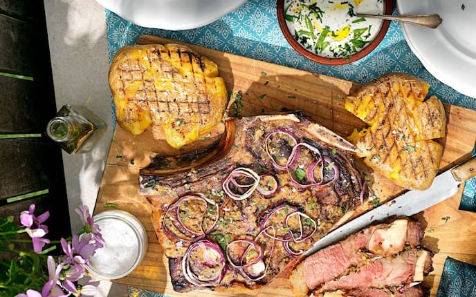 Auf einem Holzbrett liegen gegrillte, zerquetschte Erdäpfel, und ein großes Stück gegrilltes Steack mit roten Zwiebelringen. Ein Teil vom Fleisch ist aufgeschnitten, daneben liegt das Messer mit einer langen Schneide auf dem Brett. Neben dem Brett ist noch eine Schüssel mit Kräutersauce zu sehen.