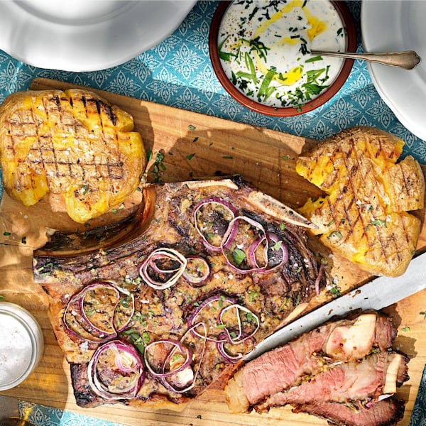 Auf einem Holzbrett liegen gegrillte, zerquetschte Erdäpfel, und ein großes Stück gegrilltes Steack mit roten Zwiebelringen. Ein Teil vom Fleisch ist aufgeschnitten, daneben liegt das Messer mit einer langen Schneide auf dem Brett. Neben dem Brett ist noch eine Schüssel mit Kräutersauce zu sehen.