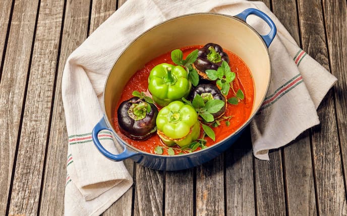 grüne und violette gefüllte Paprika in Tomatensauce in einer ovalen tiefen Rein, auf einem Holztisch mit einem Geschirrtuch rundherum