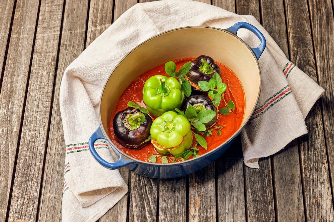 grüne und violette gefüllte Paprika in Tomatensauce in einer ovalen tiefen Rein, auf einem Holztisch mit einem Geschirrtuch rundherum