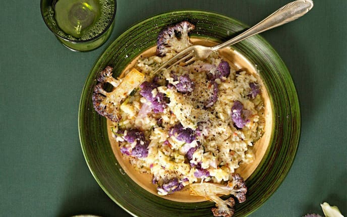 Risotto mit violetten Karfiolröschen auf einem dunkelgrünem Teller mit einem grünen Glas, das danebensteht und eine kleinen grünen Schüssel mit geriebenem Parmesan