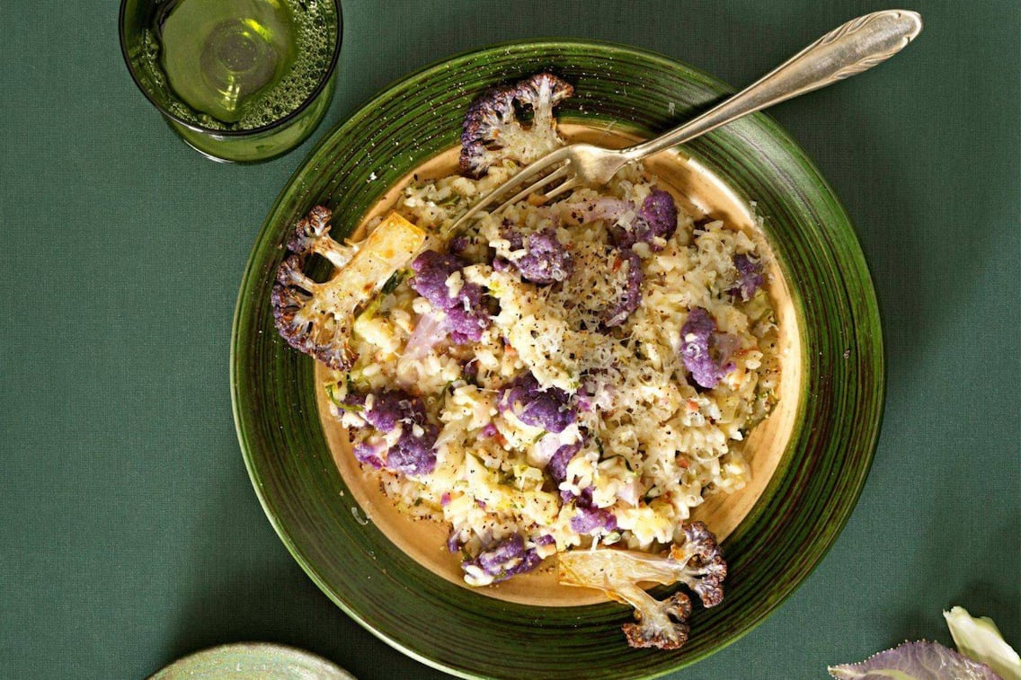 Risotto mit violetten Karfiolröschen auf einem dunkelgrünem Teller mit einem grünen Glas, das danebensteht und eine kleinen grünen Schüssel mit geriebenem Parmesan