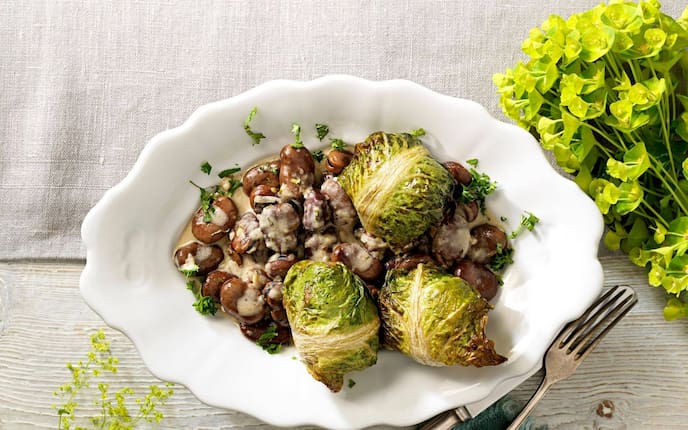 Weißer gewschungener Teller mit Saubohnen und Sauce und Kochsalatwickel. Daneben liegt ein altes Besteck.