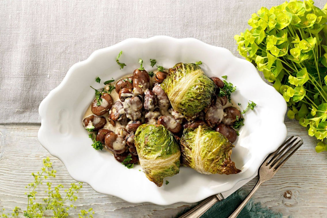 Weißer gewschungener Teller mit Saubohnen und Sauce und Kochsalatwickel. Daneben liegt ein altes Besteck.