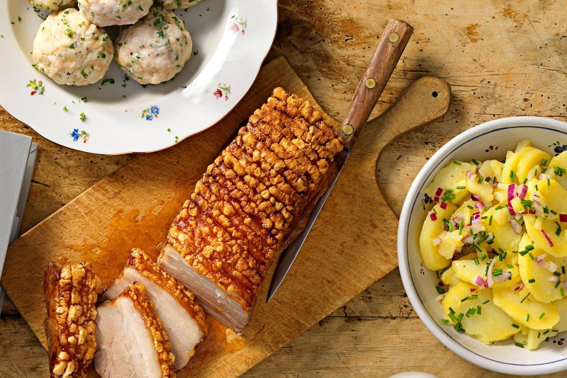 Semmelknödel auf einem geblümten tiefen Teller, Kartoffelsalat mit Schnittlauch und roten Zwiebeln, Schweinsbraten mit knuspriger Kruste auf einem alten Holzbrett, daneben liegt ein altes Messer mit Holzgriff.