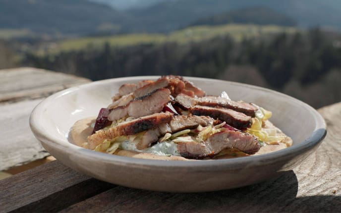 Teller steht auf einem Holztisch in Freien, im Hintergrund sind verschwommen Berge zu sehen, ein tiefer Teller mit geschnittenem Grillfleisch auf Salat mit Dressing, alles zusammen liegt auf einem Pitabrot