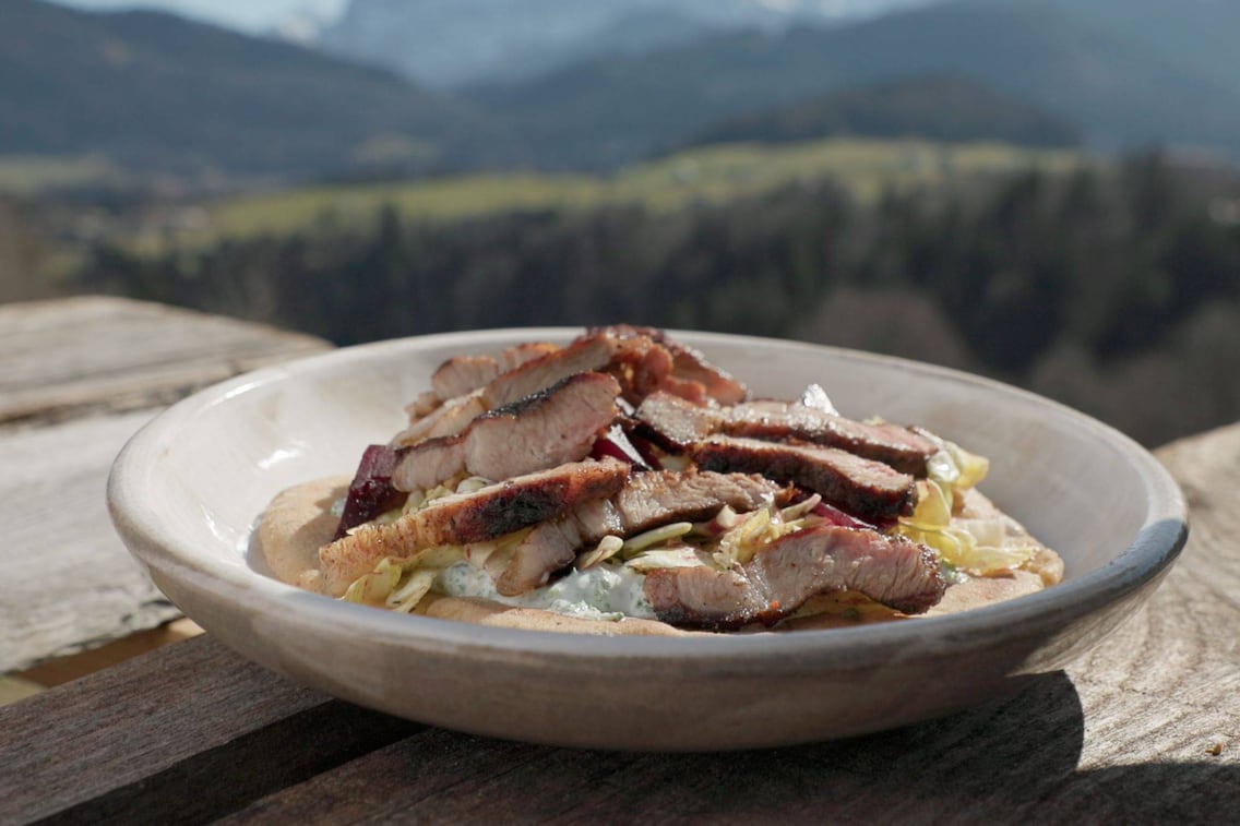 Teller steht auf einem Holztisch in Freien, im Hintergrund sind verschwommen Berge zu sehen, ein tiefer Teller mit geschnittenem Grillfleisch auf Salat mit Dressing, alles zusammen liegt auf einem Pitabrot