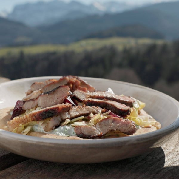 Teller steht auf einem Holztisch in Freien, im Hintergrund sind verschwommen Berge zu sehen, ein tiefer Teller mit geschnittenem Grillfleisch auf Salat mit Dressing, alles zusammen liegt auf einem Pitabrot