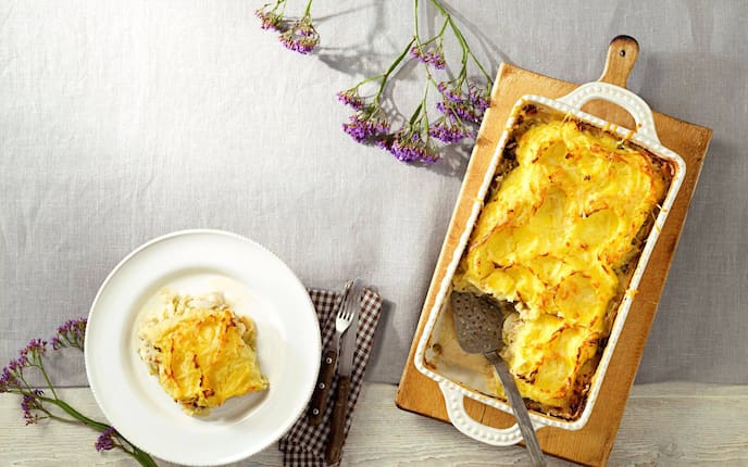 Überbackenes Frühkraut mit knuspriger Kruste in einer weißen Keramikform auf einem alten Holzbrett. Daneben steht auf einem weißen Speiseteller ein Stück von Auflauf. Daneben liegt Messer und Gabel mit Holzgriff auf einer braun karierten Stoffserviette.