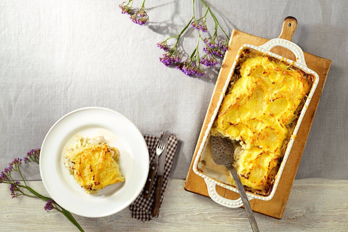 Überbackener Auflauf mit knuspriger Kruste in einer weißen Keramikform auf einem alten Holzbrett. Daneben steht auf einem weißen Speiseteller ein Stück von Auflauf. Daneben liegt Messer und Gabel mit Holzgriff auf einer braun karierten Stoffserviette.