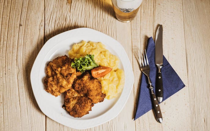Brezenschnitzel, paniert, Kartoffelsalat, Tomatenspalte, Salatblatt, Besteck, blaue Serviette, Glas Bier, Bayerische Hausmannskost, Hauptspeise, Servus Rezept