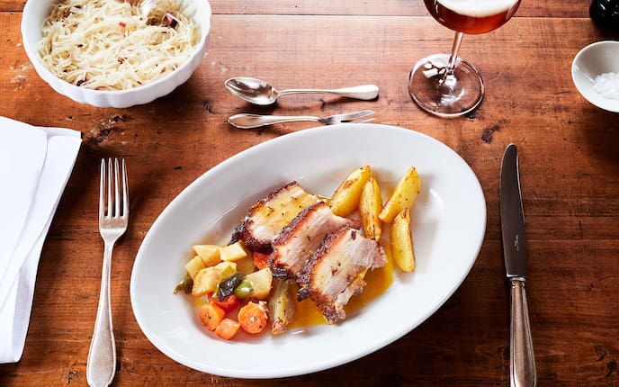 Krustenbraten vom Schwein mit warmen Krautsalat, Bier, Salz, Erdäpfelspalten, Ofengemüse, Speck, Gedeck, Holztisch