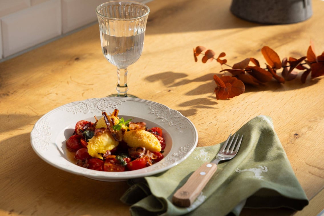 Ein weißes Teller mit Ofentomaten, Polentanockerl und Speck. Daenben eine grüße Serviette mit Hirschen drauf und ein Glas Wasser.