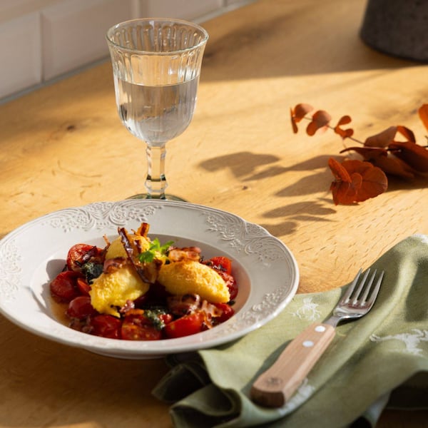 Ein weißes Teller mit Ofentomaten, Polentanockerl und Speck. Daenben eine grüße Serviette mit Hirschen drauf und ein Glas Wasser.