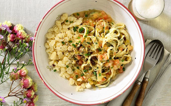 geschnittene Palatschinken mit Gemüsesauce und Spätzle in einer alten Keramikschüssel mit rotem Rand und ausgeschlagenen Ecken. Daneben liegt eine Gabel und ein Löffel mit Holzgriff auf eine naturfarbenen Stoffserviette. Daneben steht ein Glas Bier.