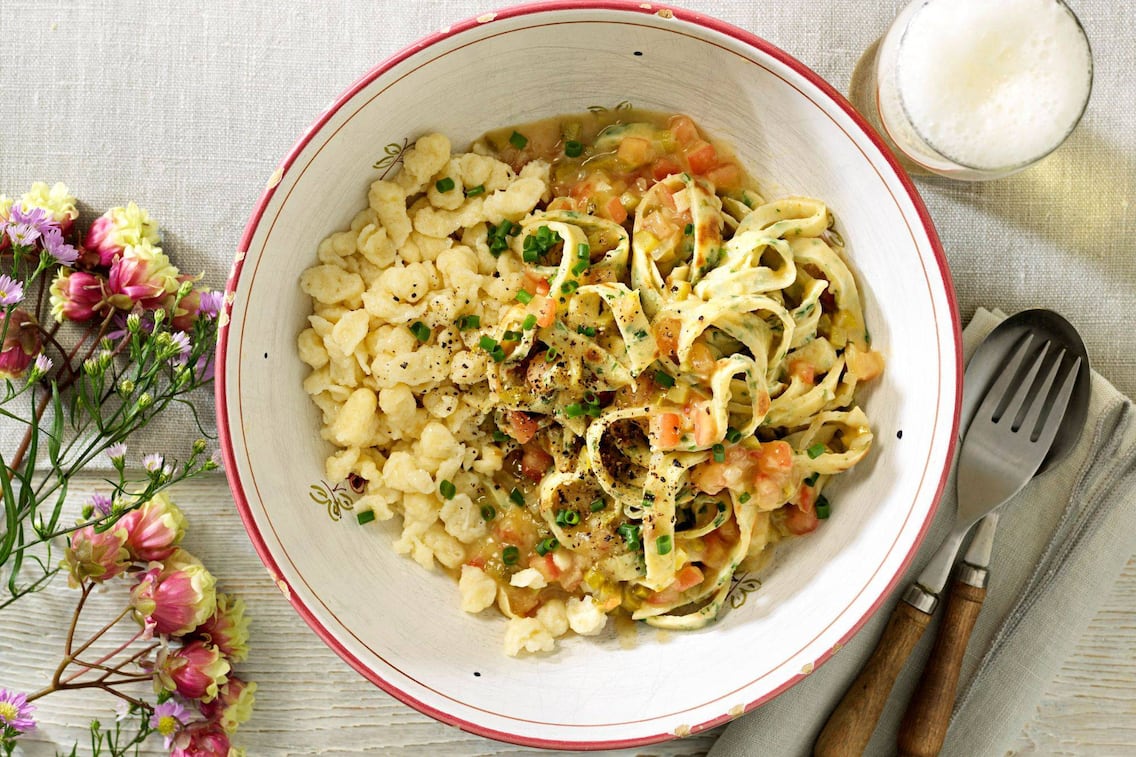 geschnittene Palatschinken mit Gemüsesauce und Spätzle in einer alten Keramikschüssel mit rotem Rand und ausgeschlagenen Ecken. Daneben liegt eine Gabel und ein Löffel mit Holzgriff auf eine naturfarbenen Stoffserviette. Daneben steht ein Glas Bier.