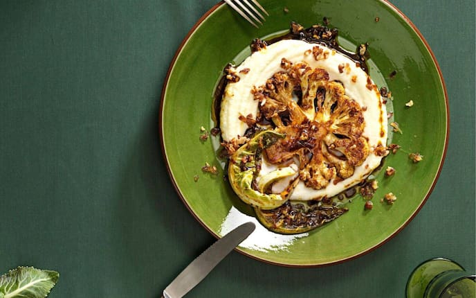 geröstete weiße Karfiolscheibe auf einem Püree mit dunkler Sauce und gebratenen Karfiolblättern. Angerichtet auf einem dunkelgrünen Teller, links ist eine Gabel, rechts vom Teller ein Messer am Tellerrand abgelegt.