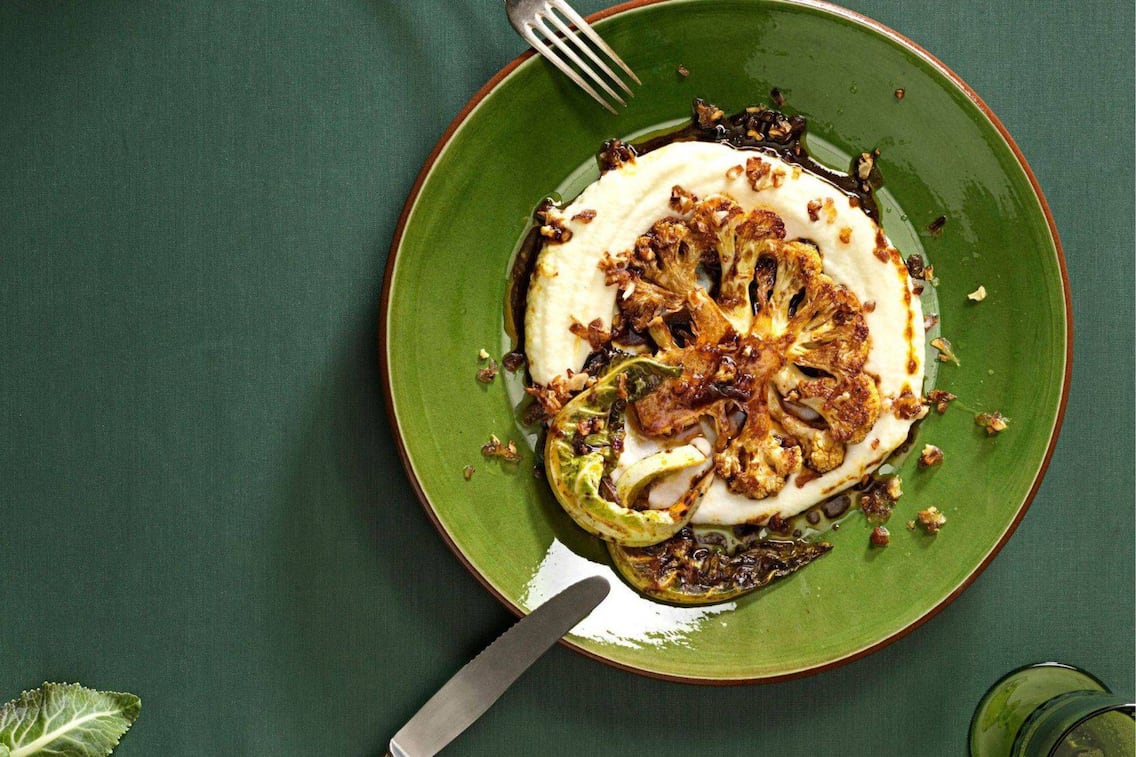 geröstete weiße Karfiolscheibe auf einem Püree mit dunkler Sauce und gebratenen Karfiolblättern. Angerichtet auf einem dunkelgrünen Teller, links ist eine Gabel, rechts vom Teller ein Messer am Tellerrand abgelegt.