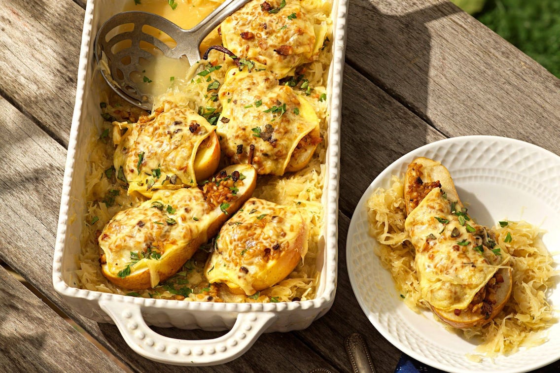 Alter Holztisch von oben fotogfrafiert auf dem eine weiße Keramik-Auflaufform steht mit gefüllten und mit Käse überbackenen Birnen- und Apfelhälften steht. Daneben ist eine Portion mit Sauerkraut auf einem Teller angerichtet.