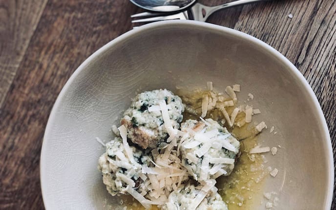 Kleine Brennnesselknöderl in einem tiefen Keramikteller mit geriebenem Parmesan und geschmolzener Butter serviert. Alles steht auf einem Holzuntergrund, daneben ligt ein Löffel und eine Gabel