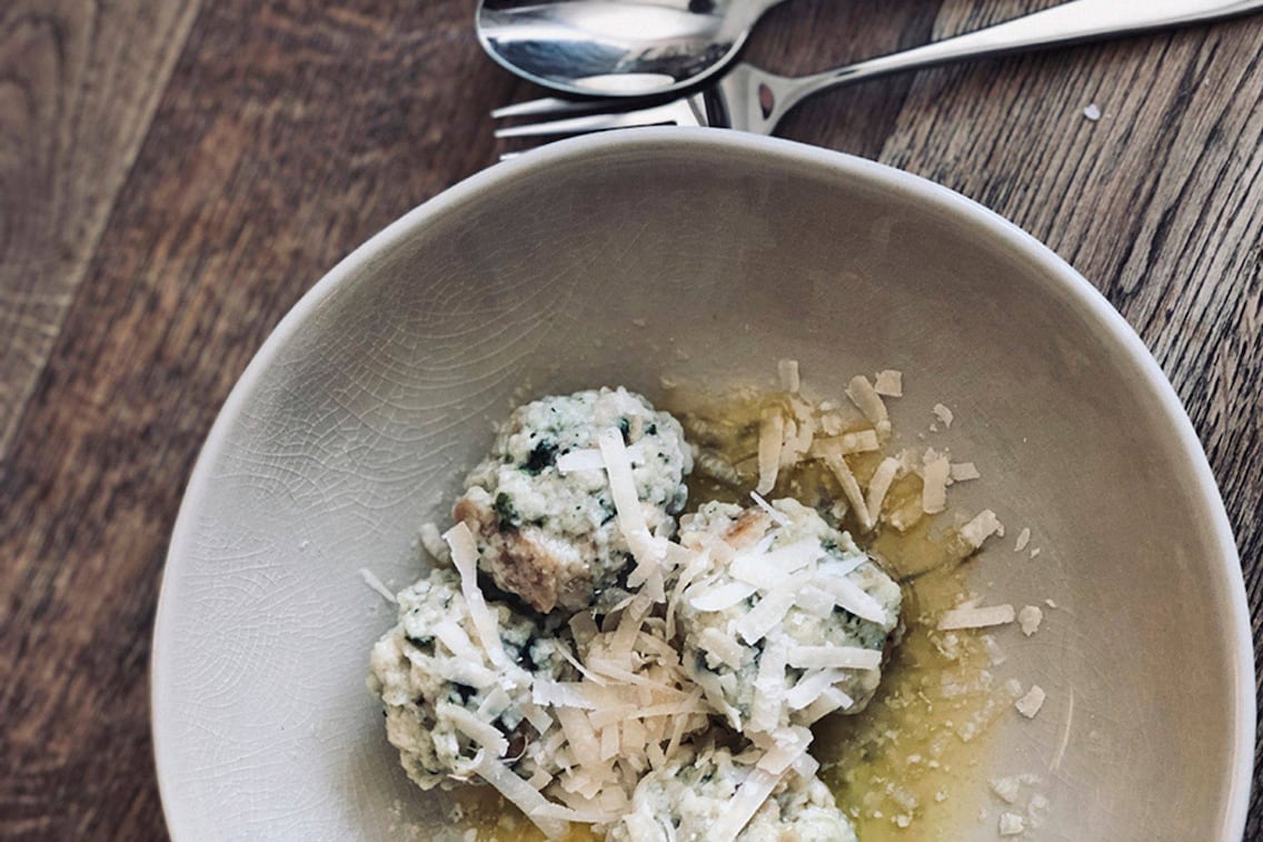 Kleine Brennnesselknöderl in einem tiefen Keramikteller mit geriebenem Parmesan und geschmolzener Butter serviert. Alles steht auf einem Holzuntergrund, daneben ligt ein Löffel und eine Gabel