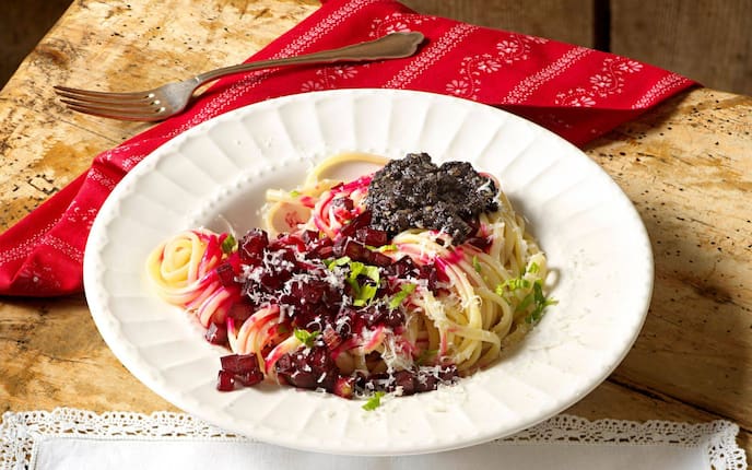 Spaghetti in einem tiefen Teller mit Rote Rüben-Würferl, Mohnpesto und geriebenen Parmesan. Daneben liegt eine rote Serviette mit weißem zarten Muster und eine Gabel. Alles steht auf einem Holzuntergrund.