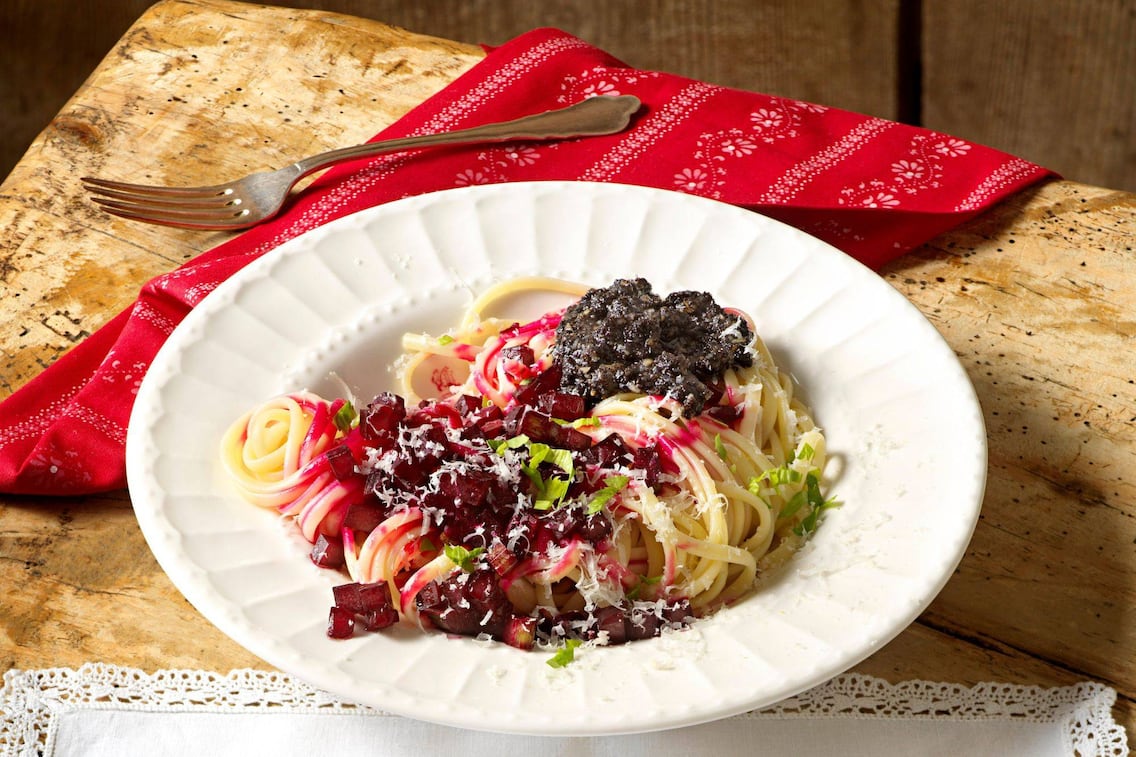 Spaghetti in einem tiefen Teller mit Rote Rüben-Würferl, Mohnpesto und geriebenen Parmesan. Daneben liegt eine rote Serviette mit weißem zarten Muster und eine Gabel. Alles steht auf einem Holzuntergrund.