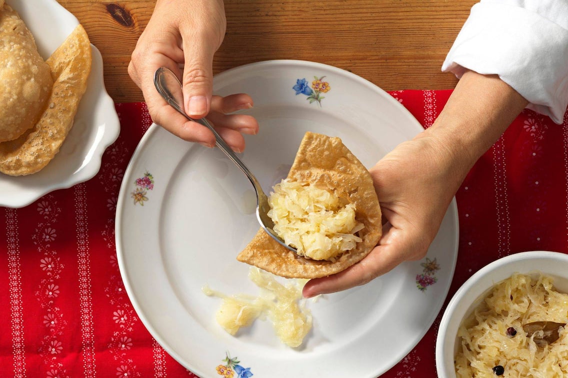 Bladln werden mit einem Löffel mit Sauerkraut belegt. Darunter steht ein weißer Keramikteller mit vereinzelten Blumenmuster. Daneben sieht man eine weiße Schüssel mit Sauerkraut und ein Servierteller mit Bladln. Alles steht auf einem Holztisch mit einer rote Tischdecke mit weißem Muster.