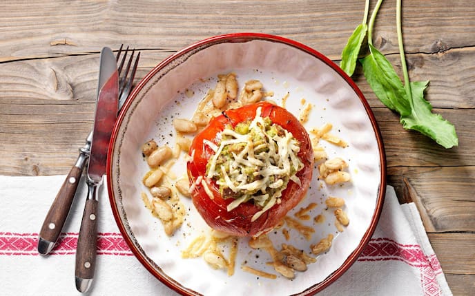 Ochsenherzparadeiser gefüllt mit einer Masse und Käse überbacken, rundherum liegen weiße Bohnen. Serviert wird es in einem tiefen Teller mit rotem Rand, danaben liegt ein Besteck mit dunkelbraunen Holzgriffen und alles zusammen steht auf einem Holztisch mit einer Tischdecke.