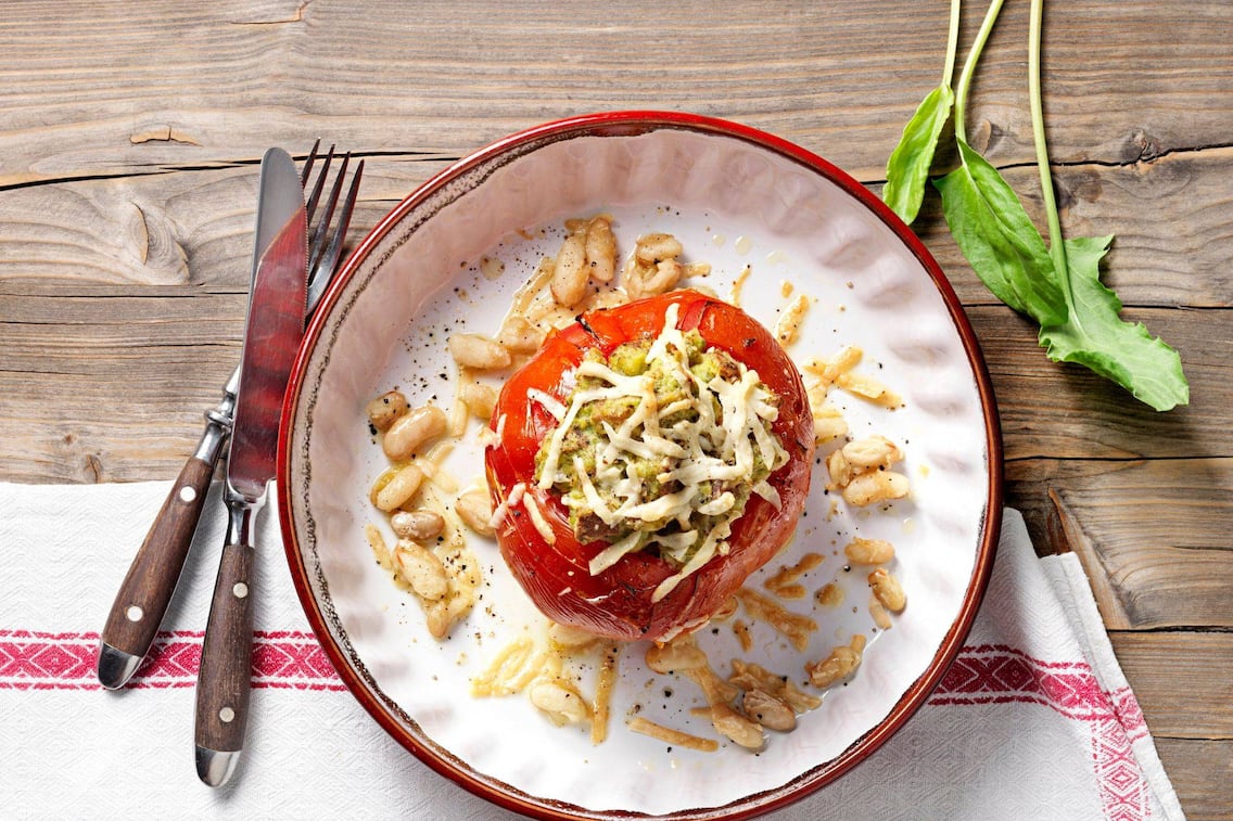 Ochsenherzparadeiser gefüllt mit einer Masse und Käse überbacken, rundherum liegen weiße Bohnen. Serviert wird es in einem tiefen Teller mit rotem Rand, danaben liegt ein Besteck mit dunkelbraunen Holzgriffen und alles zusammen steht auf einem Holztisch mit einer Tischdecke.