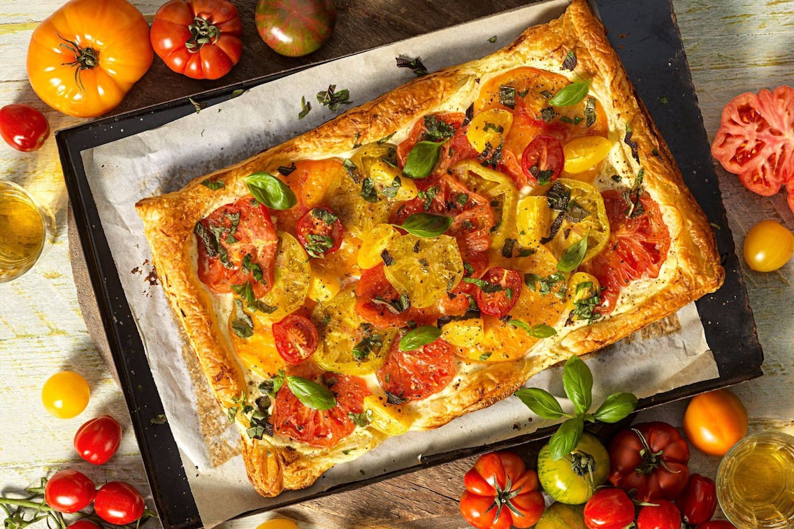 Blechkuchen mit Brimsen bestrichen und bunten Tomatenscheiben belegt. Der Kuchen liegt auf einem Stück Backpapier, das auf einem Blech liegt, das auf einem Holzbrett steht, das auf einem alten Holztisch liegt. Rundherum sind orange, rote, gelbe Tomaten verschiedener Sorten drapiert.