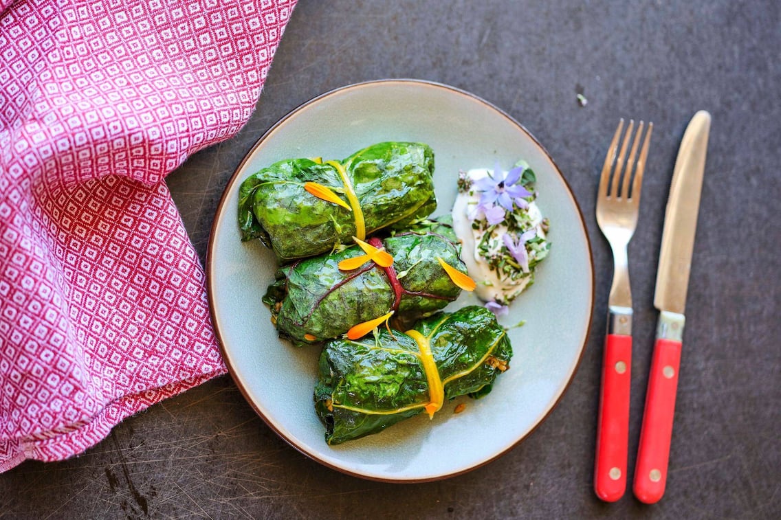 Mangoldrouladen mit einem Dip auf einem Teller angerichtet. Daneben liegt ein rotes Leinengeschirrtuch und Messer und Gabel mit einem roten Griff.