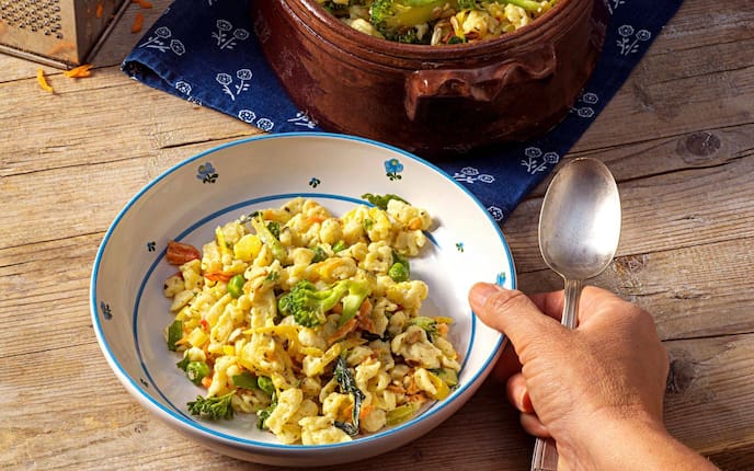 Knöpfle, Spätzle mit Brokkoli und anderem Gartengemüse verfeinert. Eine Männerhand greift mit einem Löffel zum tiefen Teller, in dem alles angerichtet ist. Der Teller steht auf einem Holztisch. Daneben steht im Anschnitt eine braune Keramikschüssel in der der Rest des Essens zu sehen ist.