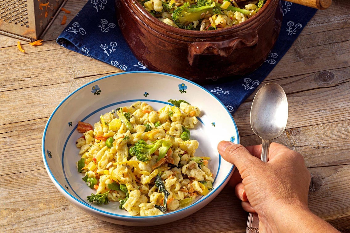 Knöpfle, Spätzle mit Brokkoli und anderem Gartengemüse verfeinert. Eine Männerhand greift mit einem Löffel zum tiefen Teller, in dem alles angerichtet ist. Der Teller steht auf einem Holztisch. Daneben steht im Anschnitt eine braune Keramikschüssel in der der Rest des Essens zu sehen ist.
